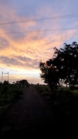 The video captures a road through a rural landscape at dusk. The sky is filled with vibrant clouds painted in shades of orange, pink, and purple against a fading blue. Silhouettes of trees and low-lying structures dot the sides of the dirt road, which stretches into the distance. The overall atmosphere is tranquil and serene, indicative of the end of the day.