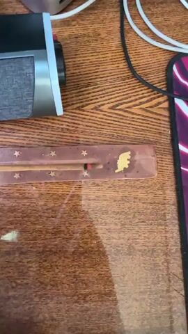 The video captures a close-up view of an incense stick burning in an incense holder on a wooden surface. The incense holder is dark brown with small gold stars and a gold silhouette of a person. The incense stick itself is thin and appears to be made of a reddish-brown material. It is lit at one end, with a glowing red ember visible. A thin wisp of smoke is rising from the burning tip. The background is blurred, but it appears to be a desk or table with some electronic devices and cables. The lighting is dim.