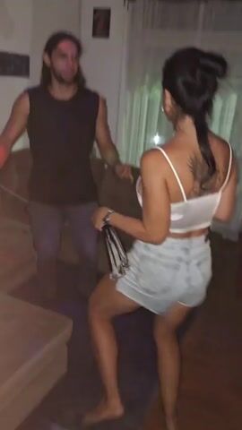 The video shows two people dancing in a living room. The man is wearing a black sleeveless shirt and jeans, while the woman has on a white top and denim shorts. Both are dancing to music with lights flashing in the background. The living room contains a couch and a table, and decorations hang on the wall. The camera is slightly shaky. The man changes into a gray t-shirt and dark pants later in the video.