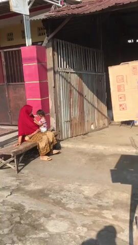 A woman wearing a red hijab and a patterned skirt sits on a wooden bench, holding a young child. The child is facing away from the camera, wearing a floral-patterned dress. They are seated in front of a large, rustic metal gate, which is partially open. To the left of the gate, there's a bright pink pillar and a dark brown metal door. Behind them, a building with a corrugated metal roof is visible. A large cardboard box with various symbols and text is leaning against the wall to the right of the gate. The ground is concrete and appears slightly wet in places, suggesting recent rain or cleaning. Sunlight casts shadows, indicating it is daytime, likely late morning or afternoon, with clear skies. The overall scene appears to be an outdoor residential or commercial area in a warm climate. The atmosphere seems calm and domestic. No specific text is clearly legible on the box or any other visible surfaces. The location is identified as Modong, Indonesia.