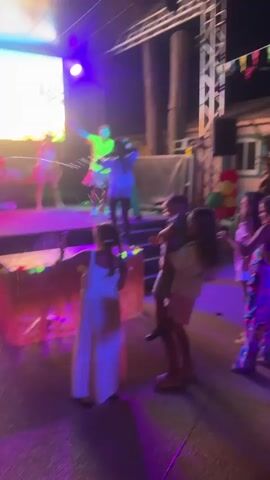 A group of people are watching a performance on a stage outdoors. There are performers on the stage dancing and singing. The stage has a large screen behind it displaying an image of palm trees and a beach. There are lights and speakers set up around the stage. In front of the stage, a group of children and adults are watching the performance. Some of the children are dancing and interacting with the performers. The environment appears to be a park or an outdoor venue, with trees and some structures visible in the background. The overall atmosphere seems to be festive and celebratory.