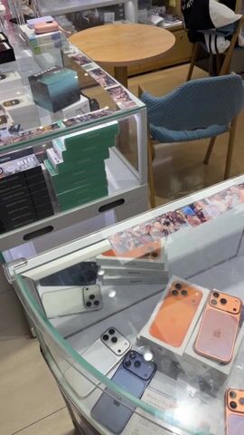 The video shows a display case filled with various models and colors of iPhones and power banks at a store that sells iPhone products. The display case is glass and the phones are in their original boxes. A person is shown scanning an item on the counter with a handheld device, and then placing the device back on the counter. There is also a small brown wallet on the counter. The surroundings include a wooden table, a blue chair, and shelves with more products. The sign in the background indicates that it is an Apple store.