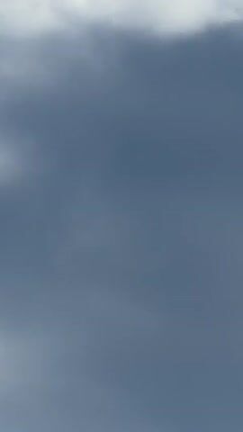 The video begins with a close-up of dark clouds, which quickly transitions to a view of a bright blue sky with scattered white clouds. A telephone pole with wires is visible in the lower left portion of the frame. The camera then pans to the right, revealing more of the sky and the telephone pole, along with a body of water and trees in the distance. The video ends with a glimpse of greenery and what appears to be the edge of a mosquito net or screen in the foreground. There are no people visible in the video.
