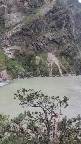 This is a video of a river flowing through a mountainous landscape. The water appears to be murky and light green. Steep, rocky hillsides covered in sparse vegetation rise on either side of the river. In the foreground, there is a bare tree with gnarled branches on the left side of the frame, and some greenery and rocks on the right. The sky is overcast and the lighting suggests it is daytime. There are no people or discernible man-made structures in this frame.