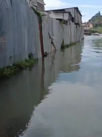 The video shows a flooded area with shacks lining one side of the water. The shacks are made of corrugated metal and appear to be in disrepair.  The water reflects the sky and surrounding trees. In the background are buildings and a line of trees along a hill.  The water level is high enough to submerge the bottom portion of the shacks.