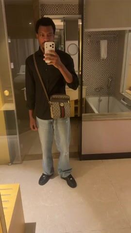 A person stands indoors, taking a photo of themself in a mirror. The individual is wearing a long-sleeved black shirt, light blue jeans, and black shoes. A tan Gucci crossbody bag is slung across their torso. The person holds a smartphone in their right hand, using it to capture the reflection. The background reveals a bathroom setting with tan tile flooring, a bathtub, and a counter visible in the mirror.  A wooden table or cabinet is visible in the left corner of the frame.