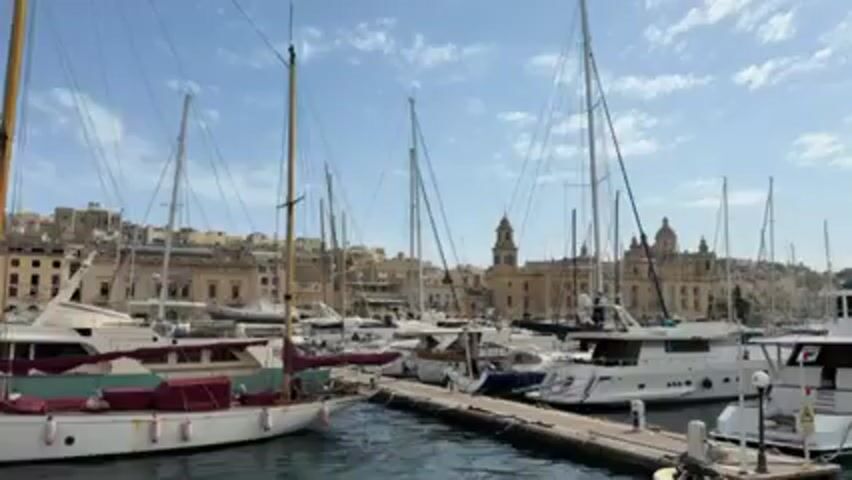 The video shows a harbor filled with various boats, including sailboats and yachts, docked along a pier. Buildings with domed roofs and clock towers are visible in the background against a partly cloudy blue sky. The water in the harbor is calm.