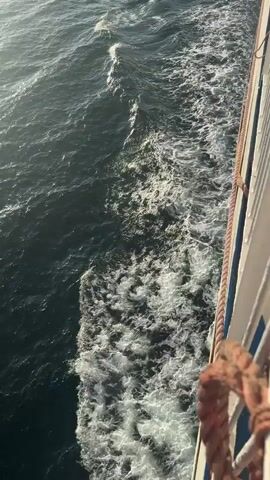 This is a vertical, POV shot from a moving ferry. The water is dark and choppy, with a white wake trailing behind the ferry on the right side of the frame. The wake is foamy and turbulent, indicating the speed of the vessel. The sun is low in the sky, casting a warm light on the water and creating a slight glare. The ferry's railing is visible along the right edge of the frame, with a thick rope tied to it. There are no people clearly visible in the frame. The horizon is visible in the distance, with a hazy sky.