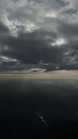 The video shows an aerial view of a landscape.  Above the landscape, there is a sky with gray, cloudy formations. The clouds are scattered and have pockets of blue sky peeking through. The sun is shining through the clouds, creating light rays in the hazy atmosphere.  Below the clouds, the ground is dark and obscured by mist or haze, and a bright line is visible winding its way through the landscape.