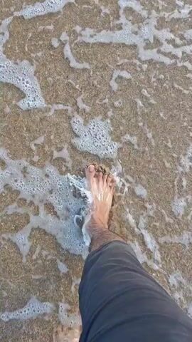 The video shows a first-person perspective of someone walking on a beach. The camera is pointed downwards, focusing on the person's bare feet and the sand and water surrounding them. The waves are gently washing over the sand and the person's feet, creating a foamy texture. The person is wearing dark pants. The overall scene suggests a relaxing day at the beach.