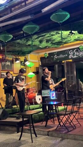 Here’s a detailed description of the video:

The video shows a live band performing on an outdoor stage in front of a bar. The band consists of four members: a keyboardist on the left, a guitarist to his right, a lead vocalist in the center, and a bass guitarist to his right as well as a drummer behind them. The vocalist stands in front of a microphone on a stand, with a music stand next to it. The band is performing on a small stage covered in artificial turf.

The background shows an Irish pub called "The Shamrock," with decorations including green lanterns and artificial foliage hanging from the ceiling. There are tables and chairs set up in front of the stage. The pub's sign mentions "St. Patrick's Day." The lighting is warm.