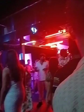 The video shows a crowded indoor space, possibly a nightclub or event venue, with a mix of people. The lighting is colorful, with red, blue, purple and green lights. There are some people standing and moving. A film crew appears to be recording in the space. There are cameras and other equipment. Some of the people appear to be part of the crew. There are others present seemingly as guests. The camera moves through the space. There appears to be a stage area in the background with some musical instruments.