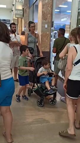 The video shows a family with two young children and a baby in a stroller in what appears to be an indoor shopping area. A man in a light-colored t-shirt and shorts is holding a baby. Beside him, a woman in a striped dress pushes a stroller with a toddler. To the left of the stroller, a young boy in a green t-shirt and blue shorts stands. Other shoppers are visible in the background, including a woman in a white dress, a man in a green shirt, and a woman in a floral top. The environment is a well-lit indoor walkway with shops on either side, indicated by signage like "TIM" and "GALLIOPI". The overall atmosphere appears casual, with people walking and shopping.