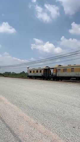 The video shows two joined railway passenger cars sitting stationary on a railway track. The cars are painted in a faded grey and gold color scheme. They appear old and possibly out of service. The cars are labeled in Thai script, with the car on the left showing the number 50818. The scene is shot in daylight under a partly cloudy sky. The ground around the track is covered with light grey gravel. Utility poles and overhead power lines are visible in the background. The camera is static.