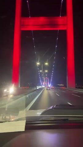 The video shows a first-person perspective of driving across a bridge at night. The bridge has multiple lanes and is illuminated by streetlights and decorative lights on the suspension cables. Red lights are visible on the large towers of the bridge. Other cars are also driving on the bridge. The road surface appears wet, suggesting it may have rained recently. The video is short and provides no additional information about the location or occupants of the vehicle.