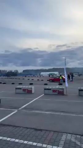 The video shows a red car drifting on an asphalt course. The car enters the scene from right and moves left, leaving tire smoke, before drifting to the right. It moves out of view, farther into the course. The course is defined by black tires. Several people are visible in the background. White concrete blocks are on either side of the frame, and say “MANNOIL FUNDACJA FUN-DRIVE DZIECIOM”. A light pole stands to the left of the frame, wrapped with striped caution tape. A stack of tires sits at the base of the pole. The sky is cloudy, and a large white building is in the background.