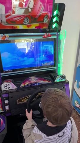 A young child with light brown hair, wearing a beige jacket and a striped long-sleeve shirt underneath, is seated in front of an arcade racing game. The child is holding a black steering wheel with both hands, positioned as if driving. The arcade machine is predominantly purple and pink, with graphics of a red sports car (resembling a Ferrari Enzo) on its upper marquee, along with some anime-style characters.

The game's screen shows a blue, cloudy sky or water effect, with what appears to be a "GAME OVER" message prominently displayed in white text in the lower-left corner. Reflections of the surrounding environment, including what looks like a window or doorway, are visible on the screen. Below the screen, the game's dashboard console features a speed indicator showing "120" and other typical racing game gauges. To the left of the steering wheel, a "START" button is visible.

The arcade machine has various lights and details, including blue arrow-shaped lights on the top marquee and green lights on the console. The child is focused on the game screen. The video is accompanied by the characteristic electronic sounds and music of an arcade game. The setting appears to be an indoor entertainment venue, possibly an arcade or a family fun center, with part of another colorful arcade machine visible on the right side of the frame.