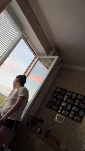A woman stands in front of a large, slanted window looking out. The window is open at the top, allowing a view of a pale blue sky with streaks of pink and orange clouds, suggesting either sunrise or sunset. The woman is facing away from the camera, her profile visible. She has dark hair pulled back in a ponytail and is wearing a white t-shirt and dark shorts with white trim. Blinds are visible on the upper part of the window. To the right of the window, a patterned wallpaper is visible, adorned with a black frame containing multiple small photos. Below the window, a dark surface, possibly a dresser or shelf, is visible with a few indistinct objects on it. The lighting in the room is dim, with the light from the window being the primary source.
