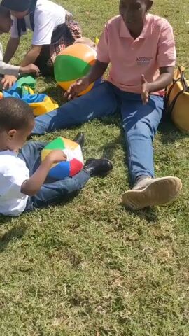 The video shows a group of people sitting on a grassy lawn, likely outdoors. Three adults and two children are visible, arranged around colorful beach balls and a multi-colored parachute or blanket. One woman, wearing a pink shirt and jeans, sits with legs outstretched, gesturing with her hands. A young boy sits near her, also with a beach ball in his lap. Another woman in a headscarf sits across from them, interacting with a young girl who is wearing a tan sweater and black pants. In the background, other people are milling around, and an inflatable bounce house is visible. The overall atmosphere seems relaxed and playful, suggesting an outdoor event or picnic.
