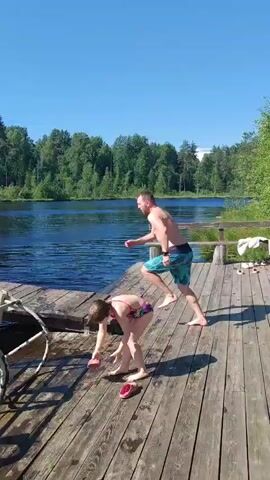 A man in blue swim trunks jumps from a wooden dock into a dark body of water. A young girl in a colorful swimsuit stands on the dock watching him. The water splashes as he hits it, creating ripples. The background shows a lush green tree line under a clear blue sky. The girl appears to be holding a pink object.