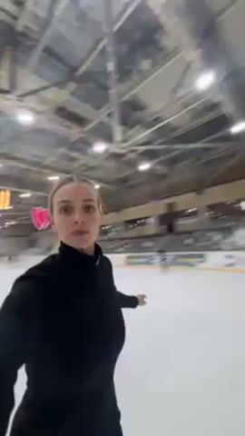 Here's a description of the video:
The video is taken from a first-person perspective, following a woman with blonde hair pulled back in a ponytail as she ice skates. She's wearing a black, form-fitting outfit. She is looking around the ice rink and smiling. The ice rink has white walls with designs, as well as glass barriers around the ice. There are other skaters visible in the background, and flags hang from the ceiling. The ceiling of the rink is metal and has hanging lights. The camera moves with the skater, capturing her movement on the ice.