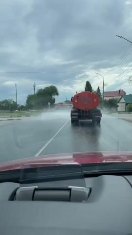 The video captures a view from inside a car, looking out through the windshield. The primary focus is a large, orange water truck driving ahead on a wet road, spraying water. The truck has the number "20" prominently displayed on its rear tank. The road appears to be in a town or village, with houses and trees visible on the sides. The sky is overcast with heavy clouds, suggesting either rain or a recent downpour. The overall atmosphere is somewhat subdued due to the weather. The time of day appears to be daytime, though the cloudy conditions might make it seem dimmer. A heads-up display unit is visible on the dashboard of the car in the foreground. The location is identified as Privolzhsky, Russia. The scene conveys the activity of street cleaning or dust suppression, indicated by the water being sprayed by the truck.
