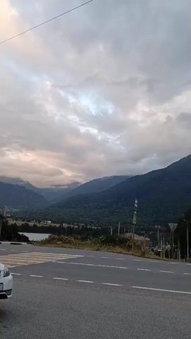 The video pans across a road in a mountainous region. The sky is overcast with clouds, and the sun is peeking through in the distance, casting a soft light on the mountains. There is a crosswalk in the foreground, and a car is visible parked on the side of the road in the distance. The landscape consists of rolling hills covered in trees and vegetation. There are also some buildings and power lines visible. The overall impression is one of a rural, scenic location.