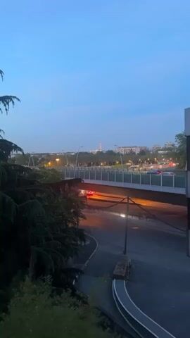 The video shows a distant view of the Eiffel Tower in Paris. The tower is lit up with colorful lights. In the foreground, there is a bridge with cars driving across it, a road with some traffic, and a line of trees. The sky is blue, and the video appears to have been taken at dusk. There's music playing in the background.