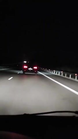 The video shows a view from inside a car at night, driving on a highway. The car in front has its brake lights on and is traveling in the same direction. The highway has white lane markers and guardrails on the right side. The overall environment is dark, indicating it is nighttime.