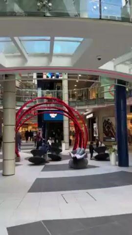 The video shows the interior of a shopping mall. The scene is centered around a seating area with several uniquely-designed, black chairs. These chairs appear to be contoured for lounging and relaxation. A large, decorative archway constructed of red, illuminated material frames the seating area. The archway is comprised of multiple curved lines. People are present in the scene, some are sitting in the chairs, lounging, and interacting with others. A child is running around the area. In the background, mall storefronts are visible, including one for Skechers. There are escalators nearby. The floor of the mall is tiled in a black and white pattern. The mall has a multi-level design with a skylight roof. The overall atmosphere is that of a modern, bustling shopping center.
