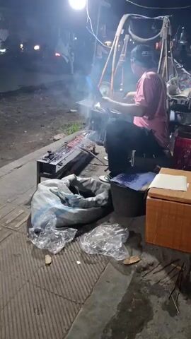 At night, a man sits near a charcoal grill, fanning the flames with a blue sheet of plastic held by a wooden handle. He wears a pink t-shirt and a baseball cap. The grill sits on a paved surface next to a large, bulging sack and a brown cardboard box. A child wearing white clothing runs past the grill.