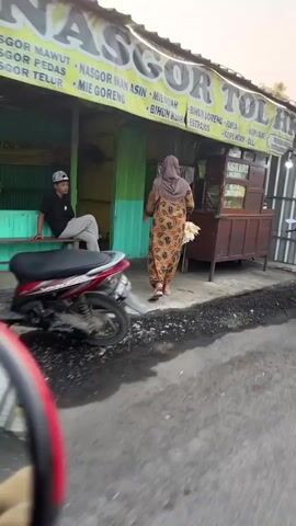 The video captures an outdoor scene at dusk. In the foreground, a motorcycle is parked on the left side of the frame. A man wearing a cap and dark clothing is seated on a green bench to the left of a food stall. The food stall has a yellow awning with text that appears to list food items such as "NASGOR MAWUT," "NASGOR PEDAS," "NASGOR TELOR," and "MIE GORENG." A woman wearing a patterned dress and headscarf is walking towards the food stall from the right. She is carrying something white in her hands. The stall itself has green and yellow accents and appears to be a simple, open-air establishment. The ground is a mixture of asphalt and gravel.