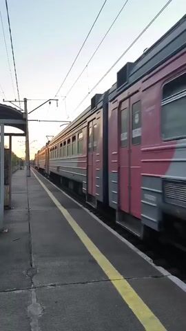 THE VIDEO SHOWS A TRAIN TRAVELING ON A TRACK ALONGSIDE A PLATFORM. THE SUN IS LOW IN THE SKY, INDICATING EITHER DAWN OR DUSK. THE TRAIN CARS ARE RED AND GRAY WITH WINDOWS. A YELLOW LINE IS MARKED ON THE EDGE OF THE PLATFORM. THERE ARE POWER LINES RUNNING ABOVE THE TRACKS. THE VIDEO IS SHOT FROM A LOW ANGLE, LOOKING ALONG THE LENGTH OF THE TRAIN.