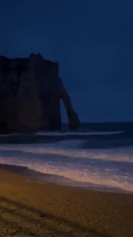 A dark, rocky arch formation stands in the ocean against a deep blue twilight sky. Waves crash onto a pebble beach in the foreground. The scene is dimly lit by artificial light, casting long shadows from an unseen source on the left side of the frame.