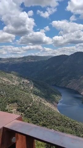 The video showcases a stunning panoramic view of a river winding through a mountainous landscape, with a partly cloudy sky overhead. Lush green forests blanket the hillsides surrounding the water, contrasting with the blue of the river and sky. In the distance, rolling hills fade into the horizon, adding depth to the scene. A bird appears to be flying in the sky. The camera is relatively still, offering a sweeping perspective of the natural beauty.