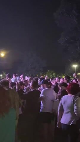 At night, a large crowd of people is shown at what appears to be a music festival, cheering as they watch a band perform on stage. The camera shifts from the crowd to the stage, where the band members are visible, playing their instruments. The environment seems to be an outdoor venue, as trees and a glowing moon are visible in the background. The stage is illuminated by lights, and the audience is enjoying the performance.