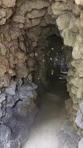 The video shows a POV walkthrough of a narrow, winding tunnel made of rough, uneven stones and rocks. The tunnel is dimly lit, with natural light filtering in through gaps and openings in the ceiling and walls, creating dappled patterns on the ground. The ground appears to be dirt and small stones. The walls are densely packed with stones of various sizes, some appearing smooth and worn, others jagged and rough. The tunnel curves and narrows as it progresses, suggesting a natural formation or an ancient, hand-constructed passage. There are no people or objects visible in the tunnel, only the stone structure and the light.