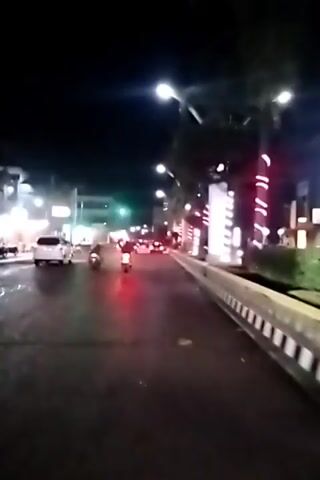 The video depicts a street scene at night. Several vehicles, including cars and motorcycles, are visible on the wet asphalt road. Streetlights illuminate the area, and buildings line the sides of the road. One building is decorated with vertical stripes of red and white lights. A white curb separates the road from a sidewalk area with trees. The traffic signal ahead is green. The video is somewhat shaky.