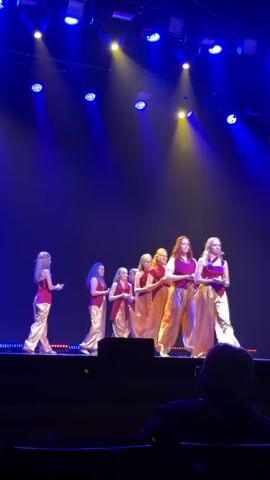 Eight young women stand in a line on a stage under bright blue and yellow spotlights. They are all wearing gold colored baggy pants and either a red or magenta colored sleeveless top. Each woman is holding a small, glowing, yellow object in front of them with both hands. The woman on the far left of the line turns to her right, away from the camera, and walks a few steps. The women are moving from left to right across the stage, with the woman on the far right being stationary while the other women are walking towards her. The camera is positioned in the audience, with a blurry dark-haired person's head visible in the foreground.