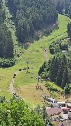 A ski lift is seen in a grassy mountain area with trees. Several cars are parked along a dirt road next to the ski lift. The ski lift towers are visible, with chairs hanging from the cables. In the background, there are more trees and a steeper, forested slope. The overall scene suggests a summer day at a ski resort, possibly with some maintenance or operational activity.