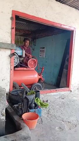 A man in a maroon shirt and dark pants is working on an orange air compressor in a garage. The air compressor has a large tank and a motor on top. The man is operating a valve on the compressor. In the foreground, there are various tools and a red bucket. The garage has a corrugated metal roof and blue walls. The entrance to the garage is framed by a red border. The ground outside the garage is concrete.