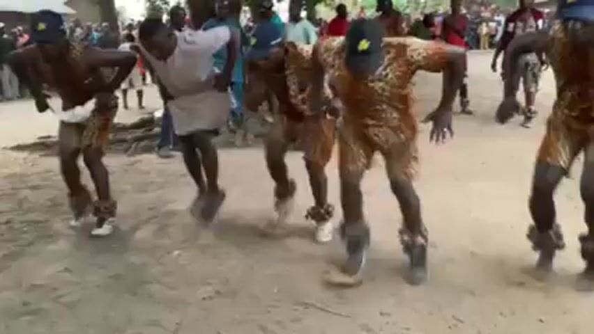 The video shows a group of men performing a dance in what appears to be a village setting. The dancers are mostly dressed in leopard-print shorts and matching hats with a leaf symbol. Some wear what seems to be a grayish tunic. They are dancing on a dirt road, kicking up dust. Most of them also have what appears to be leg rattles. A crowd of people can be seen watching in the background. The atmosphere suggests a cultural event or performance.