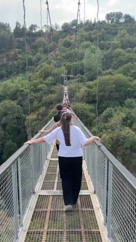 A group of people are walking across a suspension bridge. The bridge is made of metal and has a mesh walkway. The people are walking in a single file line, with the closest person to the camera wearing a white t-shirt and black pants. The bridge is surrounded by a lush green forest, with trees covering the hillsides. The sky is cloudy.