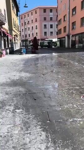 This video shows a street scene in what appears to be Italy, likely Livorno as indicated. The street is paved with stone slabs and is wet, suggesting recent rain or cleaning. Several buildings line the street, with the one on the right being a prominent peach-colored structure with multiple windows. Across the street, a lighter pink building is visible. In the middle distance, a parked green vehicle, possibly a truck or van, is present. A closed red umbrella is propped up on a cart in front of one of the establishments. The sky is clear and blue, indicating daytime. The street appears to be relatively empty, with no people clearly visible. The overall impression is one of quietness, possibly early morning or a less busy time of day in an urban environment. There is no visible text in the video.