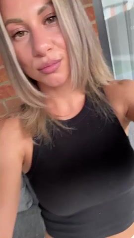 The video is a selfie video of a woman. She has blonde hair and is wearing a black crop top and white pants. She is sitting on a couch, with a brick wall behind her. She touches her hair and smiles at the camera.