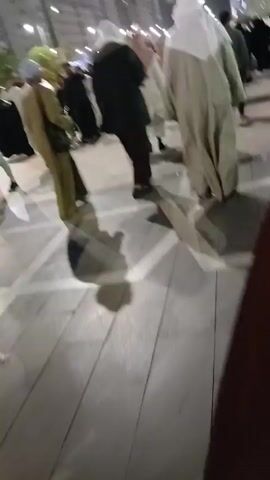 The video shows a street scene at night with a large group of people walking along a wide sidewalk. The people are wearing a variety of clothing, including traditional Muslim robes and head coverings. The sidewalk is paved with light gray bricks, and there are streetlights illuminating the scene. In the background, there are tall buildings with windows, and a bus driving by. The camera appears to be held by someone walking with the crowd.