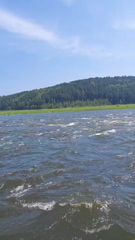 The video is a vertical shot of a fast-moving river with whitecaps under a bright blue sky with some clouds. The river is wide and choppy, with a forested hill in the background. The camera is moving forward, giving the impression of being on a boat.