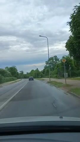 This is a first-person perspective video recorded from inside a vehicle driving on a road. The road is wet, suggesting recent rain or damp conditions. The sky is overcast with thick, gray clouds, hinting at a gloomy or unsettled weather.

The scene is characterized by a paved road with a single lane visible in the direction of travel, and another lane going the opposite direction separated by a white line. The road is flanked by green trees and vegetation on both sides, with some areas of grass and a sidewalk visible on the right. A tall, modern streetlamp stands prominently on the right side of the road. A dark-colored car is visible in the distance, ahead of the recording vehicle, driving in the same direction. Several circular manhole covers are present on the road surface.

The overall atmosphere is one of a typical drive through a somewhat rural or suburban area on a cloudy day. The time of day appears to be daytime, possibly late afternoon, given the subdued lighting. There are no visible people in the frame, aside from what can be inferred as the driver of the vehicle from which the recording is made. The video provides no audible speech or music, but the visual elements suggest a quiet, uneventful drive. No text is visible in the video.