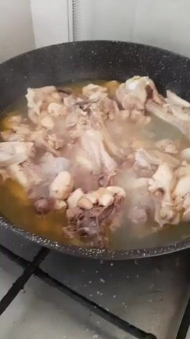 A close-up shot shows chicken pieces simmering in a dark-colored pot with a speckled interior. The chicken pieces are a mixture of bone-in cuts and smaller pieces, all appearing cooked and submerged in a light-colored broth that is gently bubbling. Steam rises from the pot, indicating that the contents are hot and actively cooking. The pot is placed on a black stovetop with visible grates, suggesting a kitchen environment. The background is out of focus, but appears to be a light-colored wall or backsplash. The lighting is neutral, illuminating the cooking process without harsh shadows.