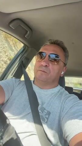 A man wearing sunglasses and a gray t-shirt is driving a car. He is looking forward and appears to be listening to something through white earbuds. The car interior is visible, including the steering wheel and a seatbelt. Outside the car, a landscape with hills and vegetation can be seen through the windows. The lighting suggests it is daytime.